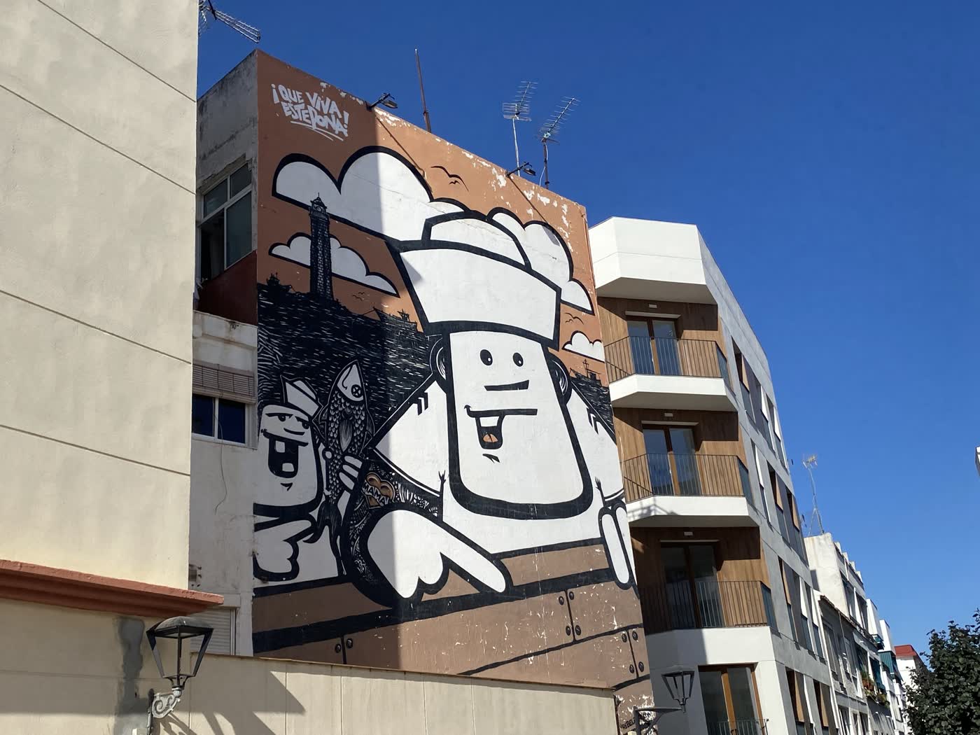 Mural «Freaks» de Rebelzer en la calle Granada de Estepona: dos personajes de dibujo vestidos de marineros con un pez, el faro y barcos al fondo, y el lema «¡Que viva Estepona!».
