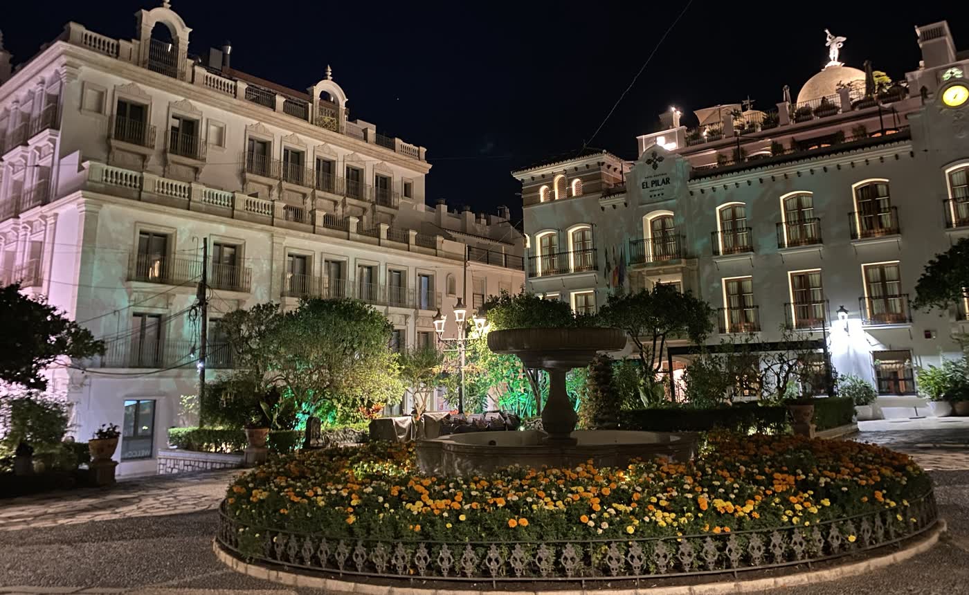 Plaza de las Flores de Estepona de noche: la fuente rodeada de flores y, al fondo, la fachada blanca iluminada del Hotel Boutique El Pilar Andalucía.