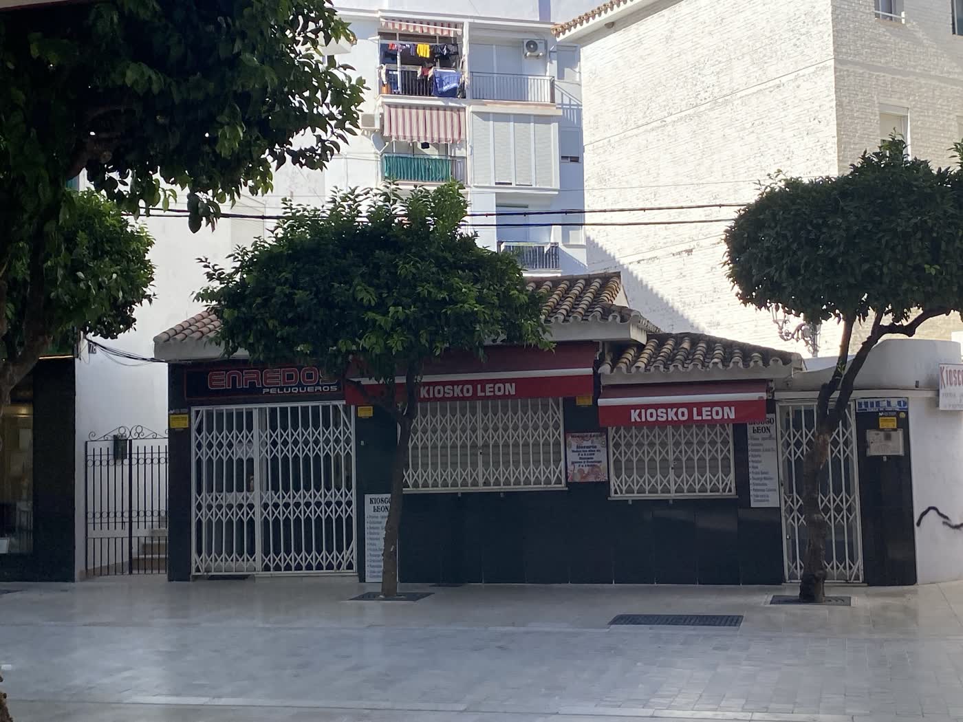 Kiosko León en la calle Terraza de Estepona: caseta con tejadillo de teja, toldos rojos con el nombre y rejas cerradas, entre naranjos y junto a la peluquería Enredos.