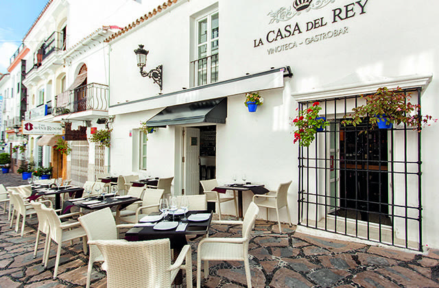 La Casa del Rey de día, en la calle Raphael de Estepona: fachada blanca con el rótulo «La Casa del Rey · Vinoteca · Gastrobar», reja con geranios y la terraza con mesas de mimbre sobre el empedrado.