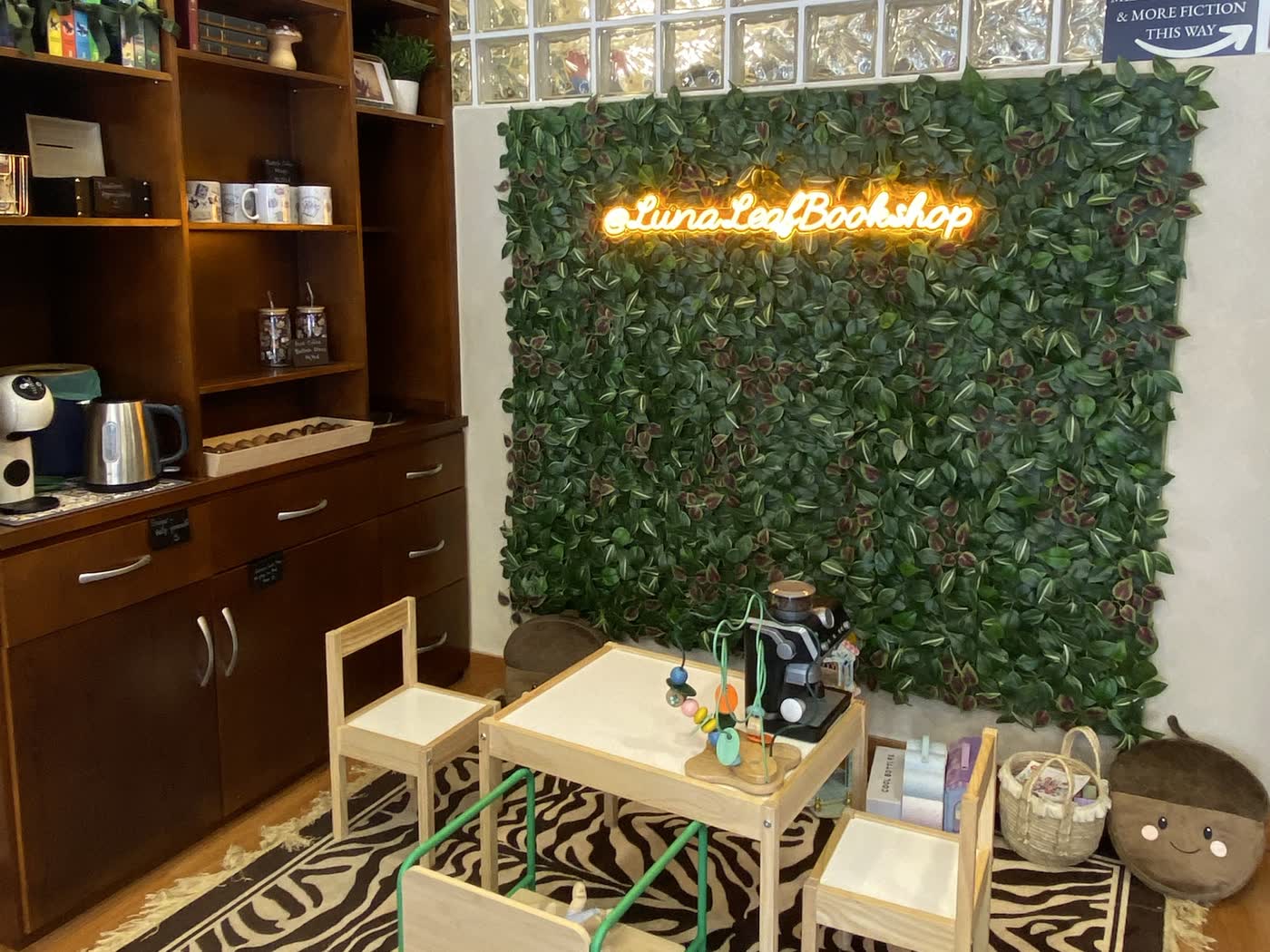 Interior de Luna & Leaf Bookshop en Estepona: rincón infantil con mesa y sillas pequeñas ante una pared de hojas con el neón «@LunaLeafBookshop», y estanterías de madera con libros y tazas.