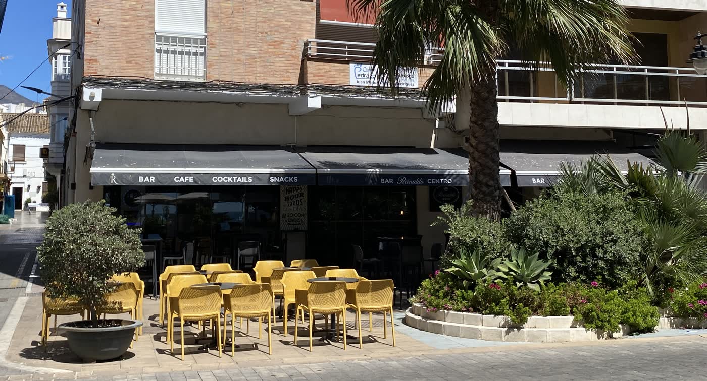 Terraza de Reinaldo Centro en Estepona: sillas amarillas bajo los toldos negros con el rótulo «Bar · Café · Cocktails · Snacks · Bar Reinaldo Centro», junto a una palmera.