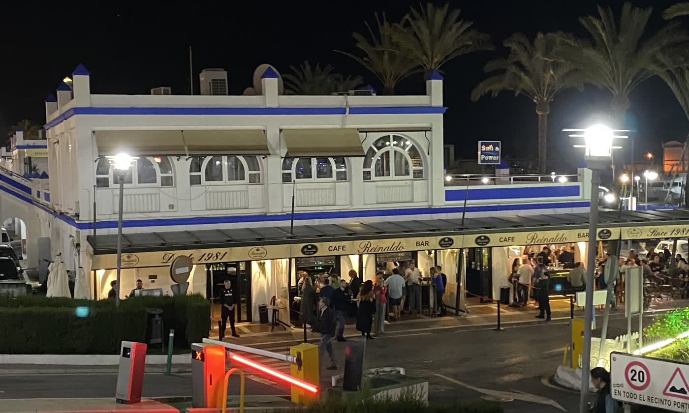 Café Bar Reinaldo en el Puerto Deportivo de Estepona de noche: edificio blanco con franjas azules, arcos iluminados y terraza llena bajo el rótulo «Desde 1981».